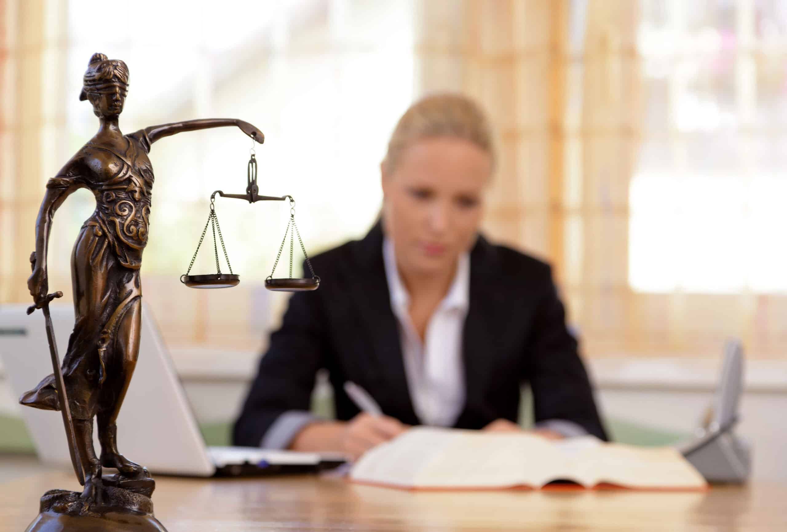 Recht Bronzefigur der Justitia mit Waage auf einem Schreibtisch vor einer schreibenden Person in Anzug